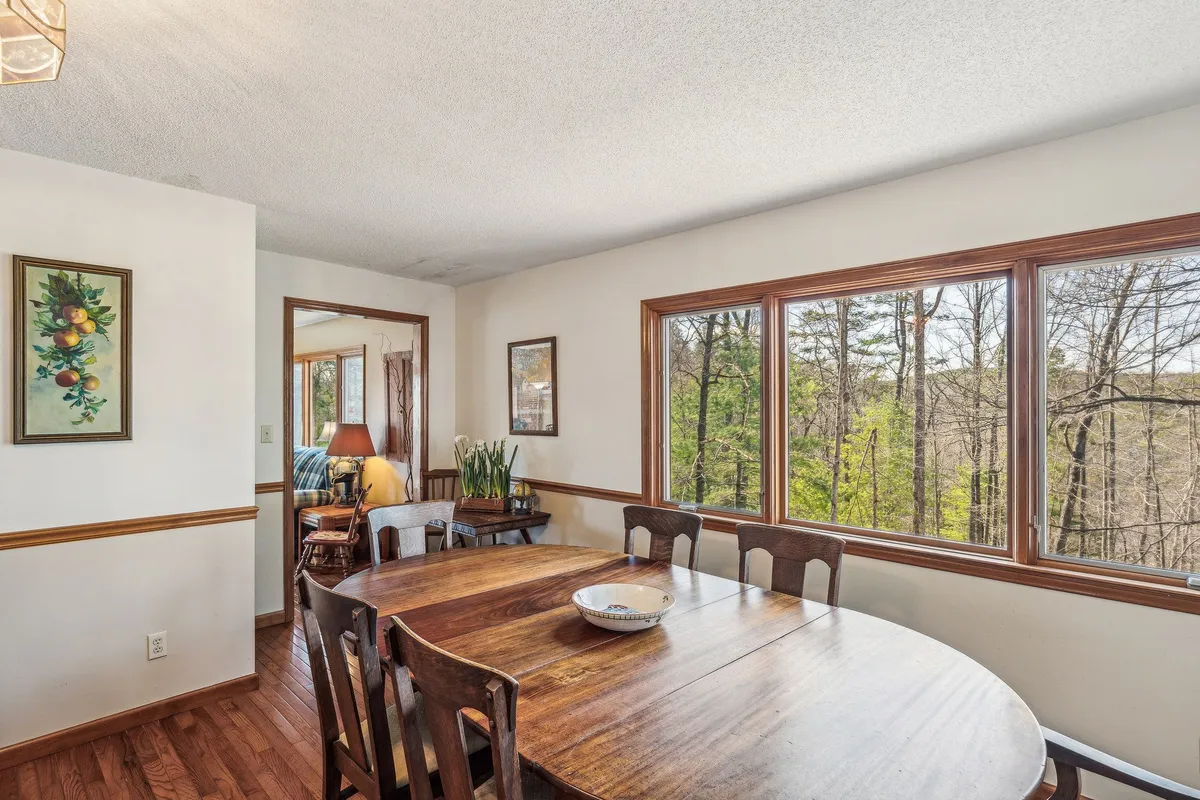 A dining area with a wooden table surrounded by chairs, large windows showcasing the natural surroundings, tasteful artwork, and lush plants. The ample daylight and greenery create a seamless transition between inside and out, fostering a calm, inviting mood.