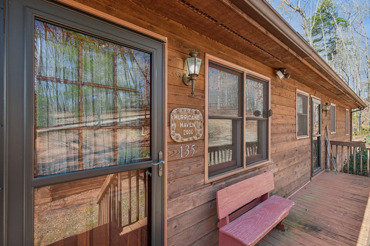 The image showcases the home's exterior front entrance with a welcoming wooden bench, house number sign, and a cozy porch area surrounded by nature. The scene feels inviting and hints at a secure, exclusive setting.