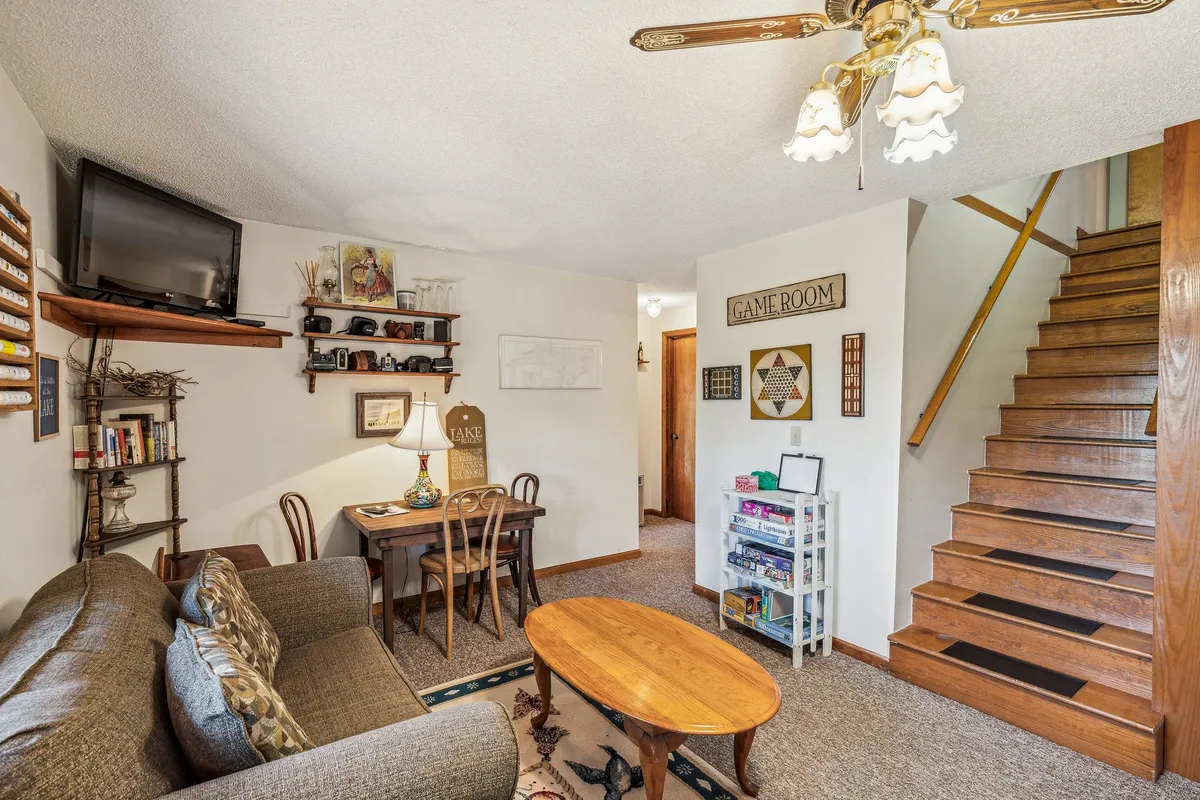 This image features a comfortable lower-level communal area with a sofa, dining table, visible staircase, and a 'game room' sign. Warm lighting, an inviting setup for gatherings, and the clear nod to games and relaxation create a playful, family-friendly atmosphere.