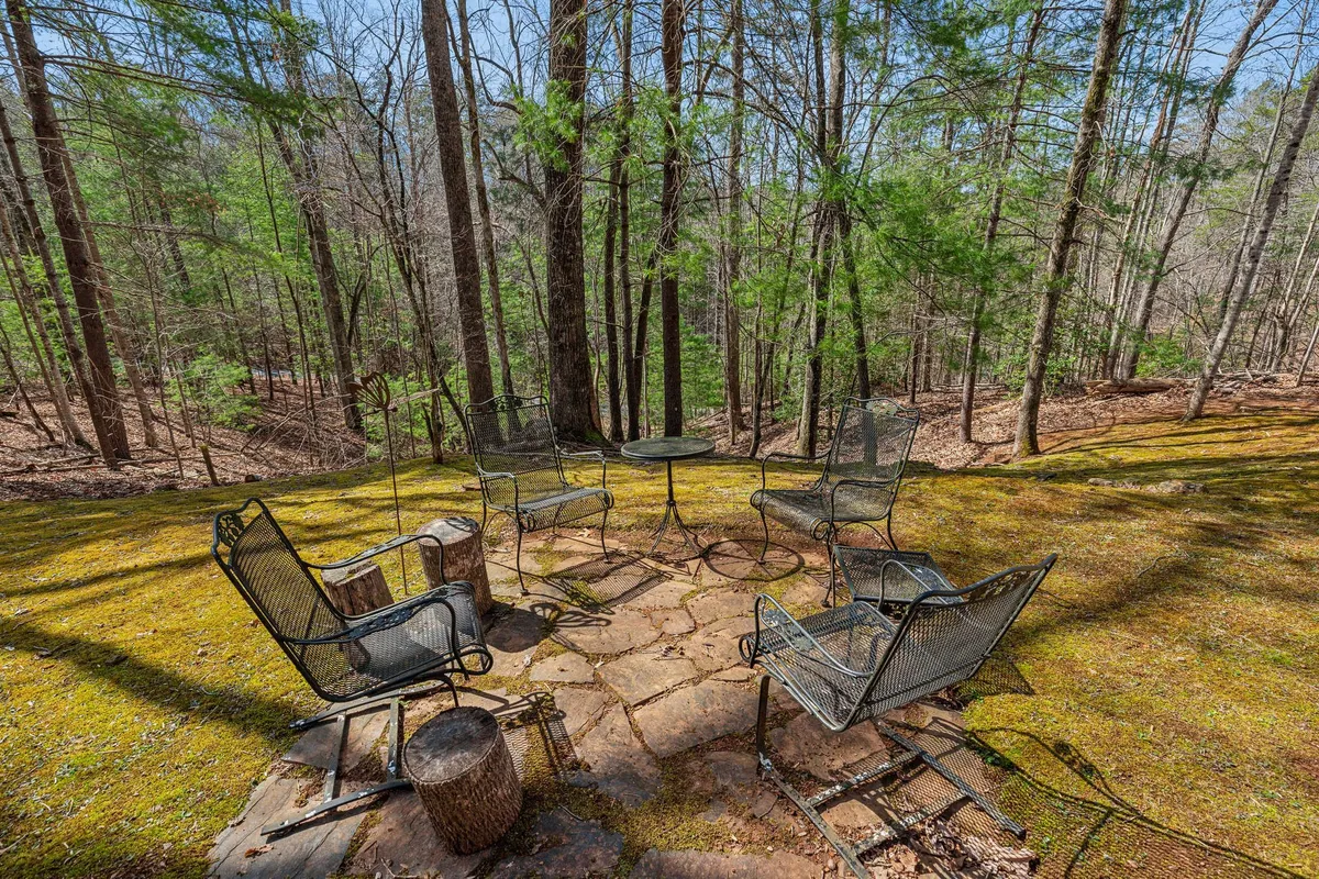 An exterior scene featuring a dining setup with metal chairs and a round table placed on mossy ground, shaded by surrounding trees. The ambiance is peaceful, suggesting opportunities for outdoor dining, casual conversations, and enjoying morning coffee while immersed in nature.