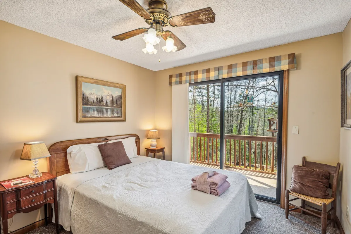 A cozy bedroom with a bed positioned to take in the view through a large window. A lamp, painting, and comfortable chair enhance the inviting ambiance, while the woodland vista outside connects the interior to the surrounding natural beauty.