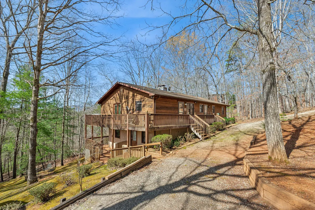The image shows a charming wooden cabin set amidst mature hardwood trees, with a welcoming deck enclosed by a railing. The house nestles peacefully in lush natural surroundings, offering a sense of seclusion and tranquility. The deck is perfectly positioned for enjoying sunsets and the soothing sights and sounds of nature.