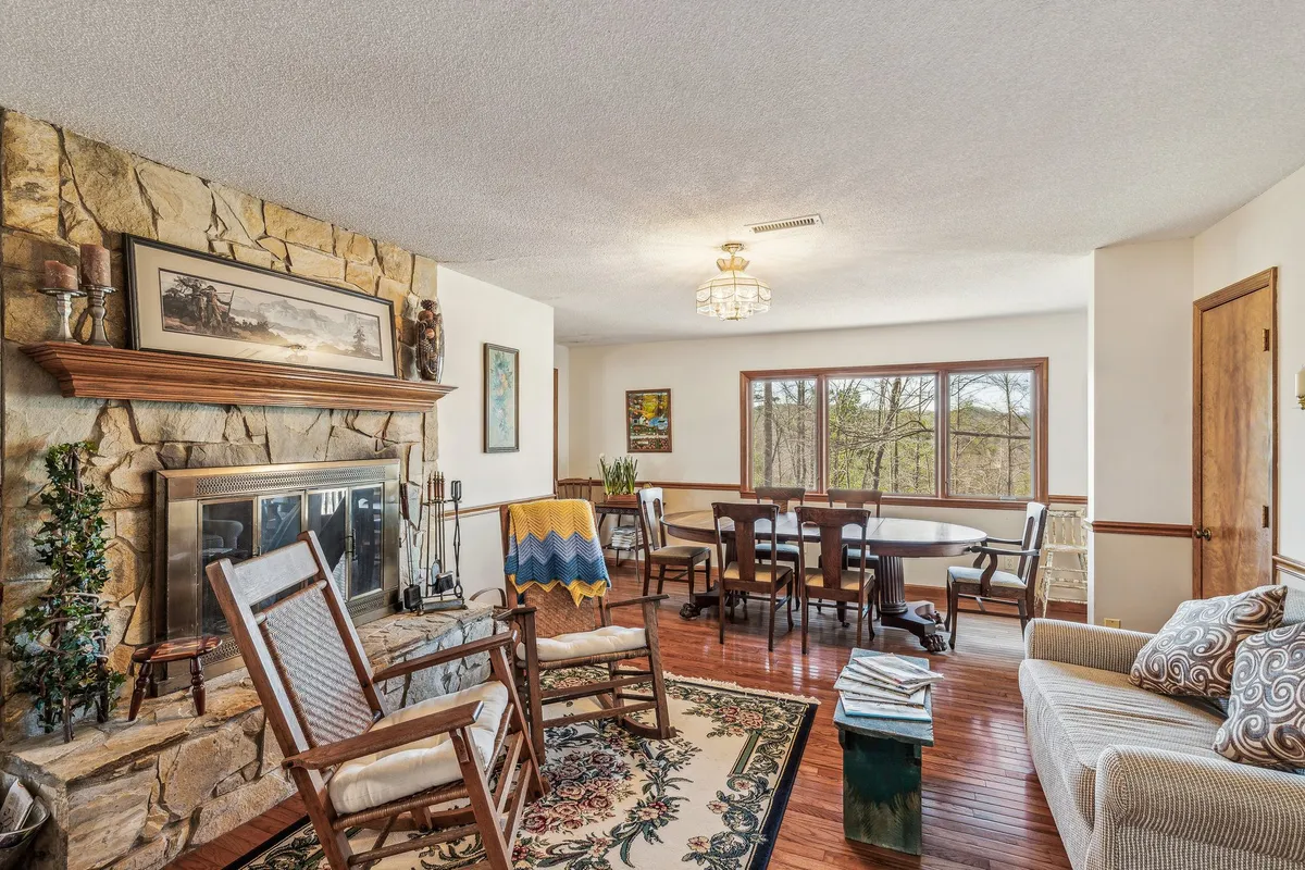 This interior image features a cozy open-plan living area with a prominent stone fireplace, a large wooden dining table, rocking chairs, and expansive windows letting in natural light. The space feels welcoming, with ample seating for gatherings and group meals.